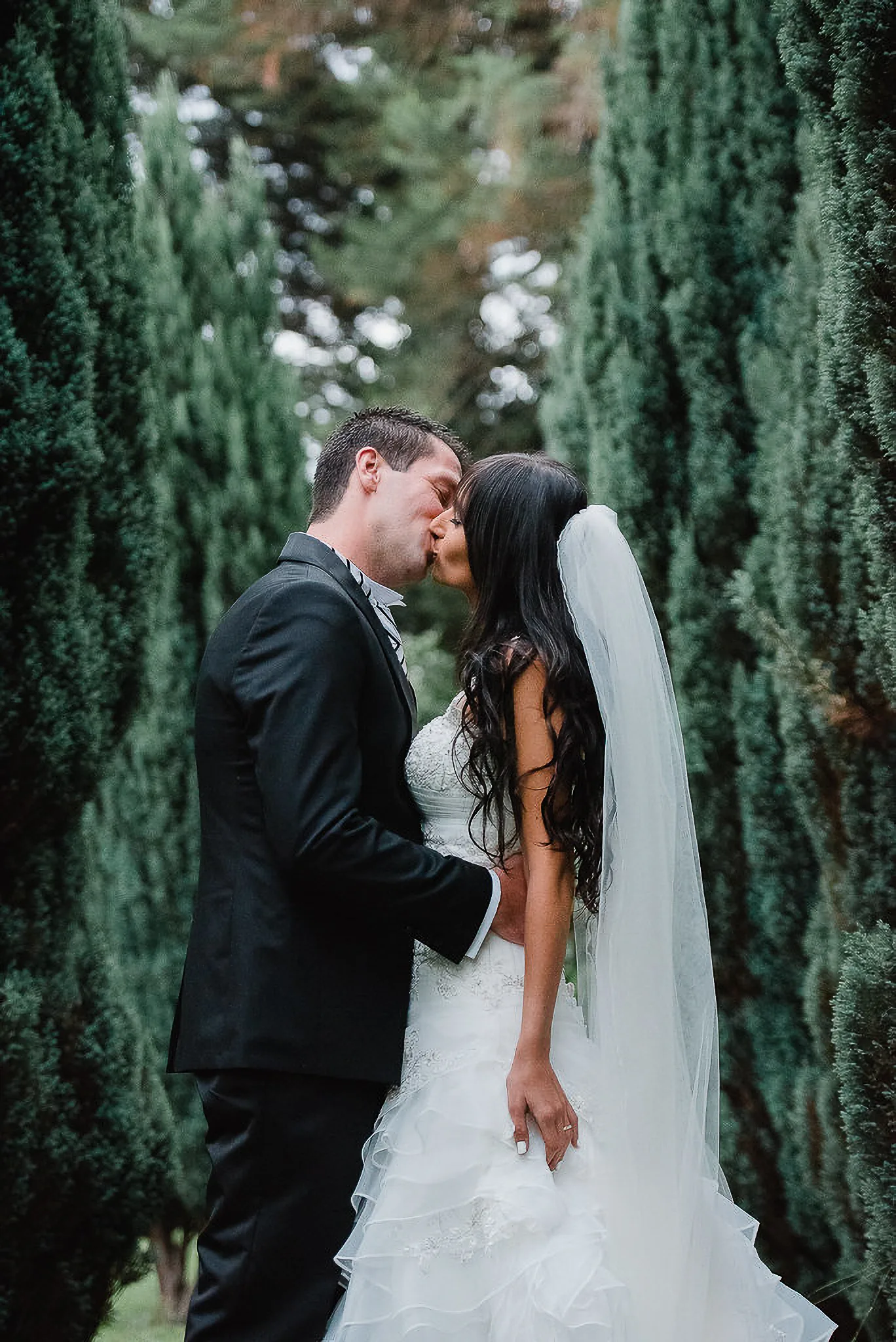 Beso de los novios entre cipreses, ella con su velo largo
