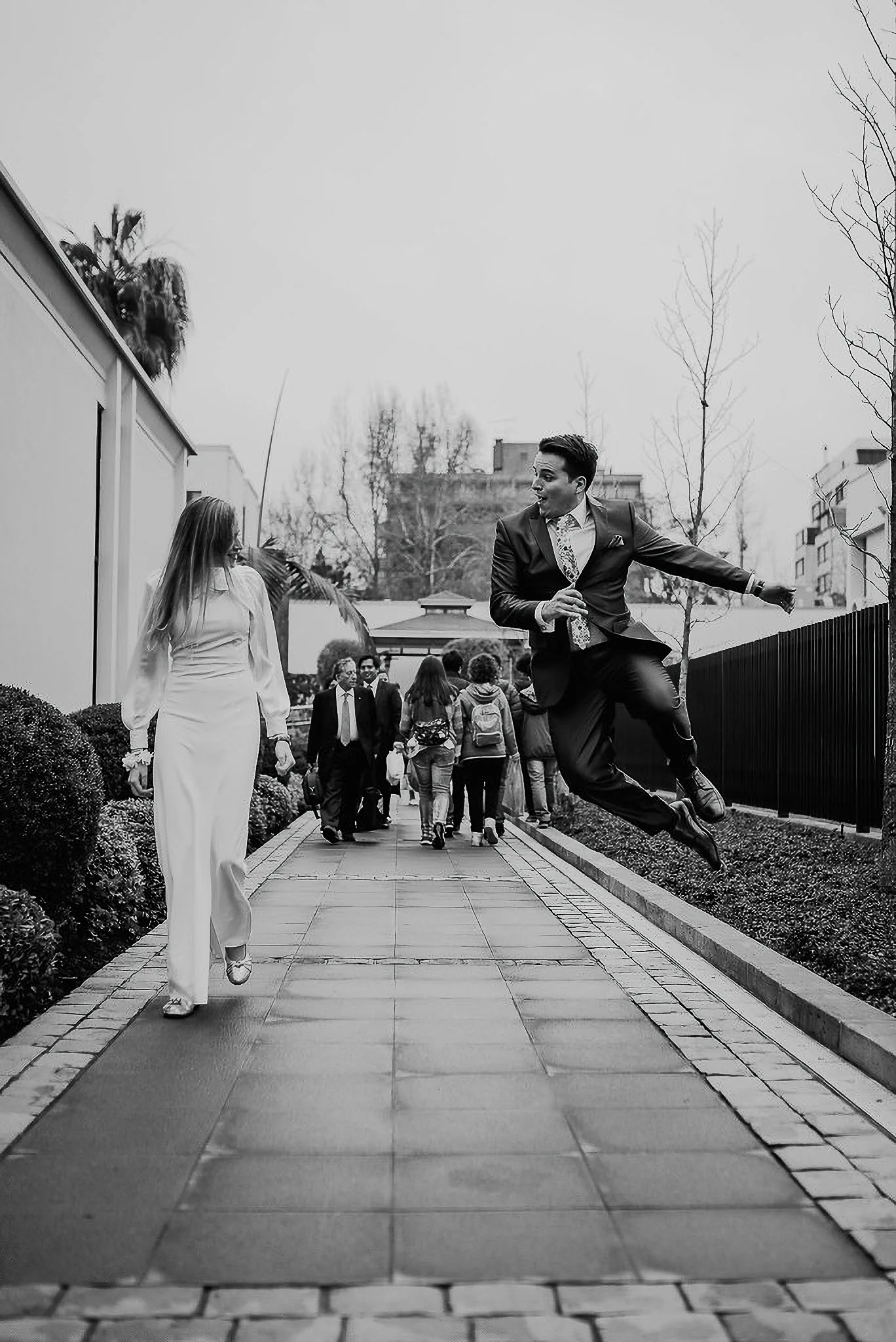 Novio saltando de alegría mientras la novia camina por el sendero, blanco y negro