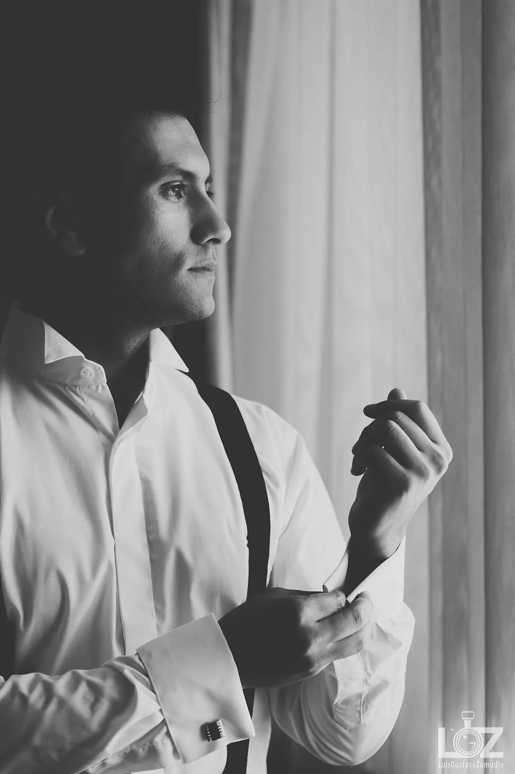 Novio abotonándose la camisa junto a la ventana durante los preparativos